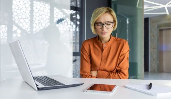 Sick woman alone at workplace has stomach cramps and period pain, sitting behind her desk in the office.