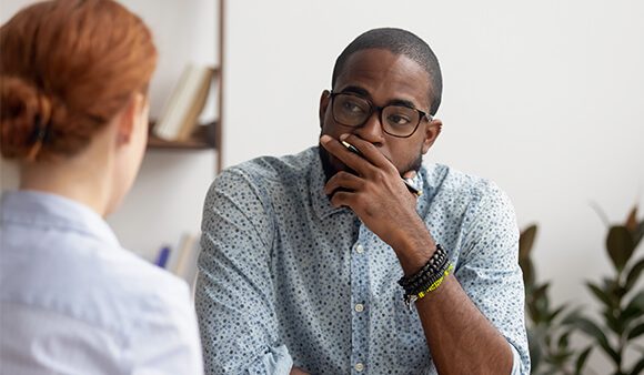 African-American man looking thoughtful and holding a pen up to his mouth whilst listening to a female colleague.