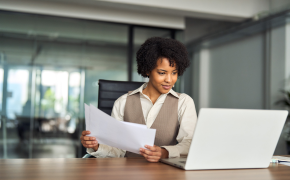 Professional busy young African American business woman account manager executive checking document financial report using laptop checking file working with enterprise software sitting at office desk.