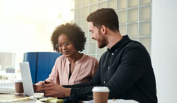 Diverse business colleagues sitting at a desk in a modern office talking together over a laptop.