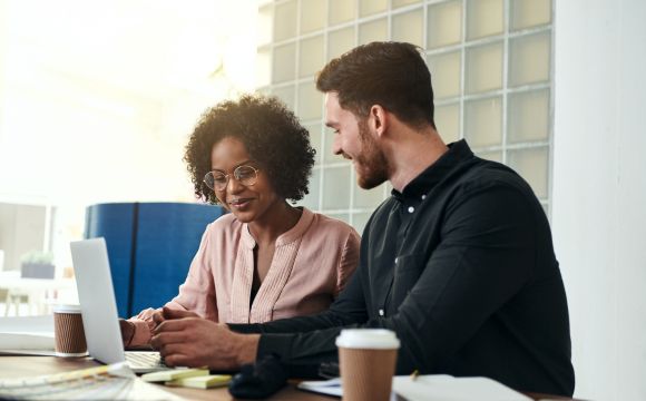 Diverse business colleagues sitting at a desk in a modern office talking together over a laptop.